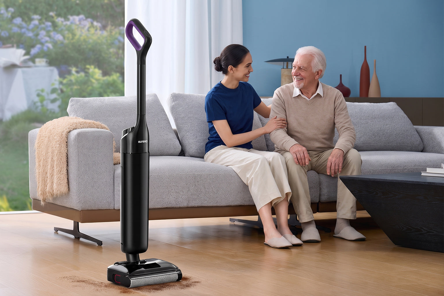 Adulto visitando a un padre anciano mientras un robot aspirador Eureka cuida la casa